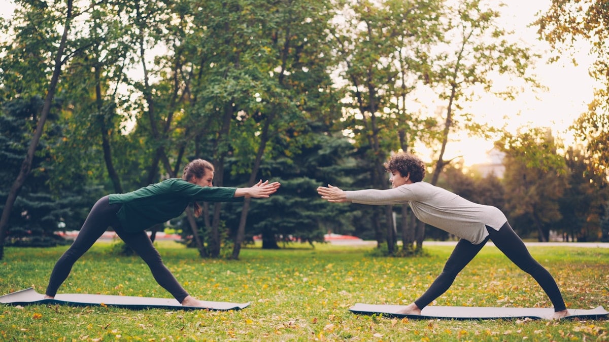 Attractive girls are enjoying outdoor yoga in city park practising positions standing on mats. Instructor is speaking teaching student, both women are relaxed and happy.